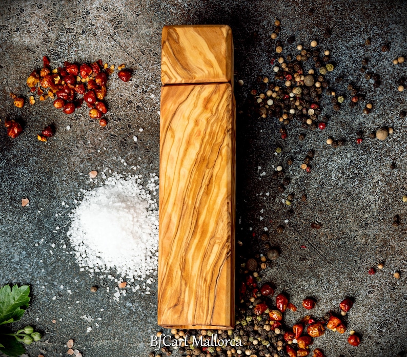 Pode incluir: Um moinho de sal e pimenta de madeira de oliveira. O moinho est&aacute; sobre um fundo cinza com gr&atilde;os de pimenta vermelha, gr&atilde;os de pimenta preta e cristais de sal branco.