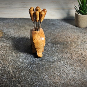 May include: A wooden appetizer pick holder with a natural wood grain finish, holding several picks with wooden handles. The holder is shaped like an elephant. The picks have stainless steel prongs. Decorative items are in the background.