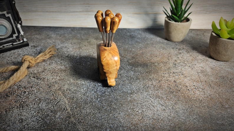 May include: A wooden appetizer pick holder with a natural wood grain finish, holding several picks with wooden handles. The holder is shaped like an elephant. The picks have stainless steel prongs. Decorative items are in the background.