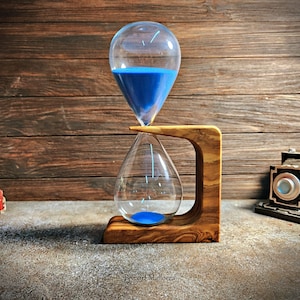 May include: A blue hourglass with a wooden stand. The hourglass is made of glass and has a blue liquid inside. The stand is made of olive wood and has a unique design.