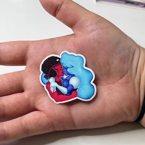 May include: A colorful sticker featuring two cartoon characters embracing. One character has red skin and black hair, while the other is blue with light blue hair. The sticker is held in a hand.