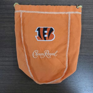May include: Orange drawstring bag with the Cincinnati Bengals logo and the "Crown Royal" script. The bag has a stitched border and a drawstring closure. The logo features the letters "B" and "C" in black and orange.