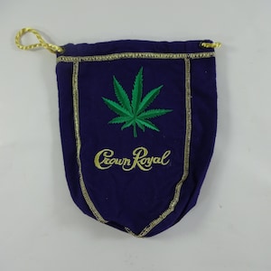 May include: A purple drawstring bag with a gold trim and a green marijuana leaf embroidered on the front. The bag also has the words "Crown Royal" embroidered in gold.