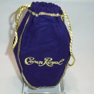 May include: A deep purple drawstring bag with gold trim and a gold drawstring. The bag features the embroidered text "Crown Royal" in gold script. The bag is made of a soft, velvety material.