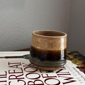 May include: A handmade ceramic tumbler with a speckled tan top, transitioning to a dark brown and black base. The tumbler sits on a book titled "The Great Book of Wine". The book's cover is white with red text.