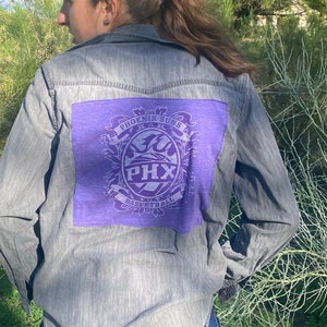 May include: A gray denim shirt with a purple patch on the back. The patch features a basketball logo with the text "Phoenix Suns" and "PHX Basketball".