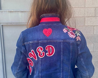 May include: A blue denim jacket with red and white embroidered details. The jacket has a red heart, the word "Loved" in red letters, and red roses embroidered on the back.