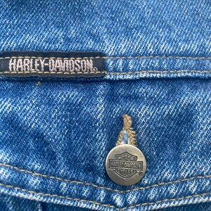 Peut inclure: Gros plan sur une veste en jean bleu avec un patch en cuir noir qui indique "Harley-Davidson" et un bouton en m&eacute;tal argent&eacute; avec le logo Harley-Davidson.