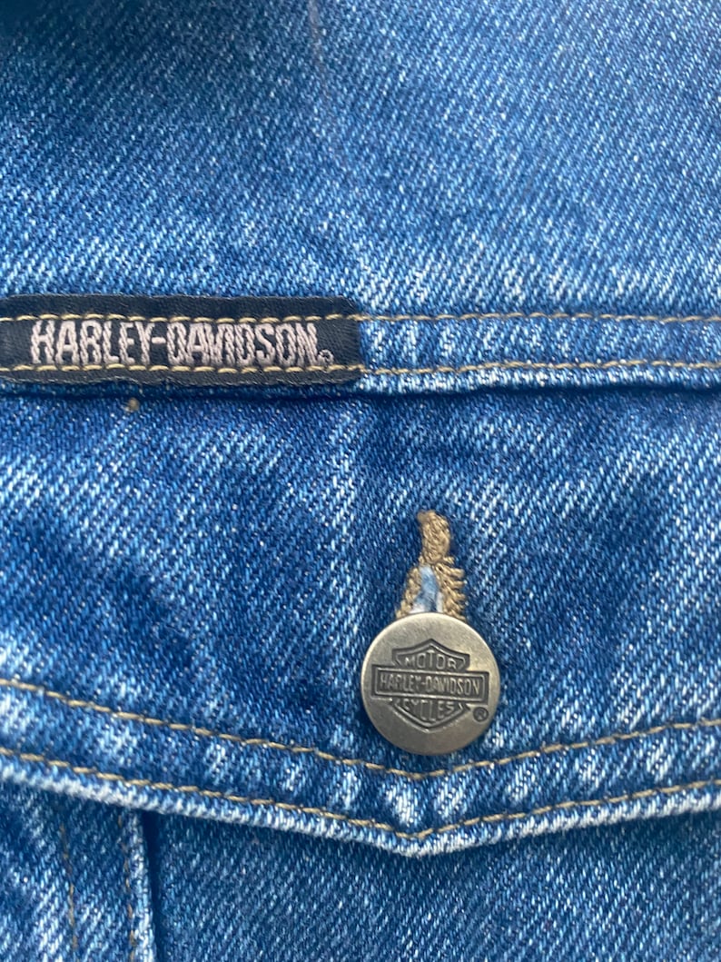 Peut inclure: Gros plan sur une veste en jean bleu avec un patch en cuir noir qui indique "Harley-Davidson" et un bouton en m&eacute;tal argent&eacute; avec le logo Harley-Davidson.