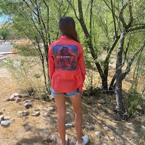 May include: A person wearing a coral button-down shirt with a "OneRepublic" album cover printed on the back. The person is wearing denim shorts and white sneakers. The person is standing in a wooded area with rocks on the ground.