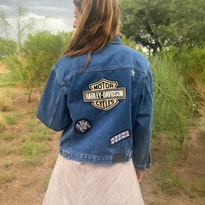 Peut inclure: Une veste en jean bleue avec un patch logo Harley-Davidson au dos. La veste a &eacute;galement d'autres patchs, dont un qui dit "Live the life you love, love the life you live".
