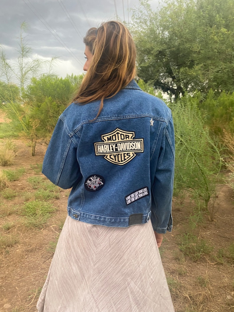 Peut inclure: Une veste en jean bleue avec un patch logo Harley-Davidson au dos. La veste a &eacute;galement d'autres patchs, dont un qui dit "Live the life you love, love the life you live".