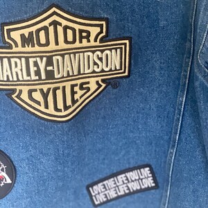 Peut inclure: Une veste en jean bleue avec un patch logo Harley-Davidson, une &eacute;toile argent&eacute;e et un patch noir et blanc qui dit "Love the life you live".