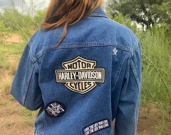 May include: A blue denim jacket with a Harley-Davidson patch on the back. The jacket also has a star patch and a black and white patch with a skull and crossbones. The text on the jacket reads "Love the life you live, Live the life you love."