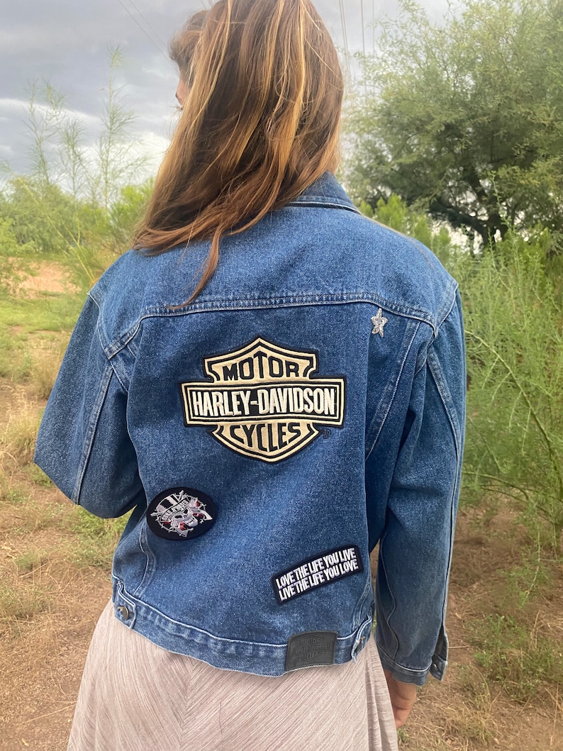 Peut inclure: Une veste en jean bleue avec un patch Harley-Davidson au dos. La veste a &eacute;galement un patch &eacute;toile et un patch noir et blanc avec un cr&acirc;ne et des os crois&eacute;s. Le texte sur la veste dit "Love the life you live, Live the life you love."