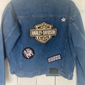 Peut inclure: Une veste en jean bleue avec plusieurs patchs, dont un logo Harley-Davidson, un cr&acirc;ne avec un chapeau haut de forme et un patch de texte qui dit "LOVE THE LIFE YOU LIVE LIVE THE LIFE YOU LOVE".