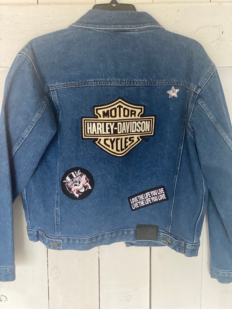 Peut inclure: Une veste en jean bleue avec plusieurs patchs, dont un logo Harley-Davidson, un cr&acirc;ne avec un chapeau haut de forme et un patch de texte qui dit "LOVE THE LIFE YOU LIVE LIVE THE LIFE YOU LOVE".