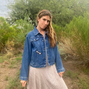 Peut inclure: Une femme portant une veste en jean bleue avec une &eacute;pingle "FRIEND" noire et blanche sur le c&ocirc;t&eacute; gauche de la poitrine. Elle porte une robe marron clair et a de longs cheveux bruns.