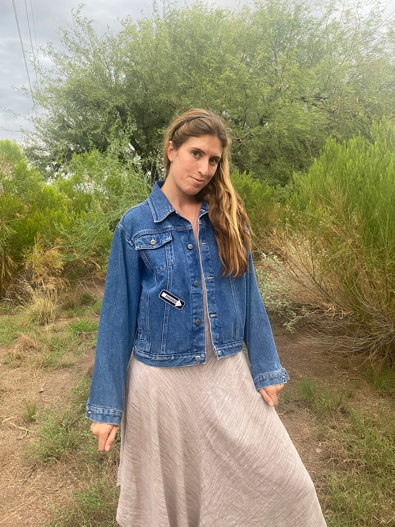Peut inclure: Une femme portant une veste en jean bleue avec une &eacute;pingle "FRIEND" noire et blanche sur le c&ocirc;t&eacute; gauche de la poitrine. Elle porte une robe marron clair et a de longs cheveux bruns.