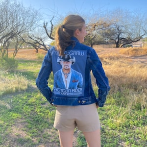 May include: A blue denim jacket with a Tim McGraw Shotgun Rider Tour 2015 patch on the back. The patch features a photo of Tim McGraw wearing a cowboy hat and a blue shirt.