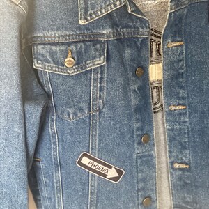 Peut inclure: Une veste en jean bleue avec un patch noir et blanc qui dit "PHOENIX" avec une fl&egrave;che pointant vers la droite.