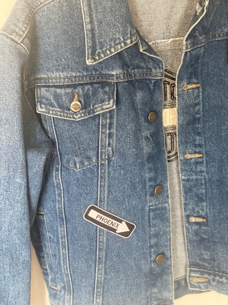 Peut inclure: Une veste en jean bleue avec un patch noir et blanc qui dit "PHOENIX" avec une fl&egrave;che pointant vers la droite.