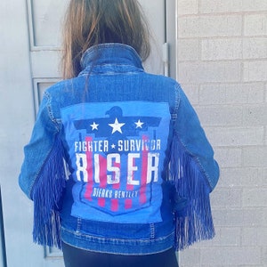 Puede incluir: Una chaqueta vaquera azul con detalles de flecos en las mangas. La parte posterior de la chaqueta presenta un parche azul y rojo con el texto "Fighter * Survivor Riser Dierks Bentley".