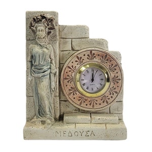 May include: A decorative clock with a white face and black hands, set in a stone-like frame with a sculpted figure of Medusa. The clock face is surrounded by a decorative border with gold accents. The base of the clock is inscribed with the word "MEDOUSA".