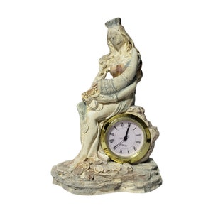 May include: A white ceramic figurine of a woman sitting on a rock with a gold clock face. The clock face has Roman numerals and the word "QUARTZ" printed on it.