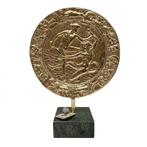 May include: A gold-coloured, circular decorative plaque depicting figures in relief, mounted on a green marble base. The plaque features a detailed design with multiple figures and a decorative border. The base is a dark green, rectangular block.