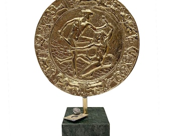 May include: A gold-colored, circular decorative plaque depicting figures in relief, mounted on a green marble base. The plaque features a detailed design with multiple figures and a decorative border. The base is a dark green, rectangular block.