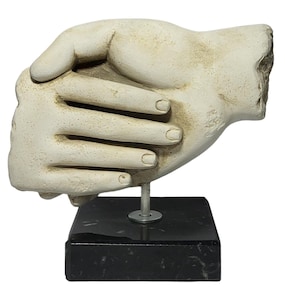 May include: A cream-colored sculpture of two hands clasped together, mounted on a black marble base. The hands are detailed with visible knuckles and fingernails. The sculpture is supported by a thin metal rod.