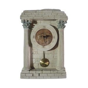 Puede incluir: Un reloj blanco de piedra con péndulo. La esfera del reloj tiene números romanos y una imagen de un edificio. El reloj está enmarcado por dos columnas.