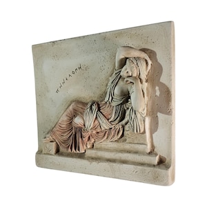 May include: A rectangular, off-white relief sculpture depicting a reclining figure in classical attire. The figure is draped in flowing robes, with one arm supporting the head. The Greek word "&Pi;&Eta;&Nu;&Epsilon;&Lambda;&Omicron;&Pi;&Eta;" is inscribed on the upper left. The sculpture has a weathered appearance.