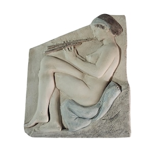 Op de afbeelding: Een lichtgekleurde reliëfsculptuur van een zittende figuur die een fluit bespeelt. De figuur is naakt, met een hoofdbedekking en een blauwe doek onder zich. De sculptuur is gemonteerd op een rechthoekige, getextureerde achtergrond.