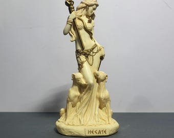 May include: Ivory-colored statue of a woman holding a staff with a flame, adorned with gold-colored jewelry. Two dogs sit at her feet. The base of the statue has the word "HEKATE" inscribed. The statue is approximately 10 inches tall.