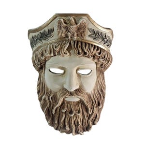 Może przedstawiać: Kremowa maska przedstawiająca brodatą twarz z koroną. Maska ma szczegółowy wzór, w tym wydatny nos, zamknięte oczy i długą, płynącą brodę. Korona przedstawia orła i liście laurowe.