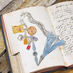 May include: A silver dragon bookmark with a blue tassel and charms, including a bag of chips, a TV, a bottle of soda, and a remote control. The bookmark is on a page of a book with handwritten text.