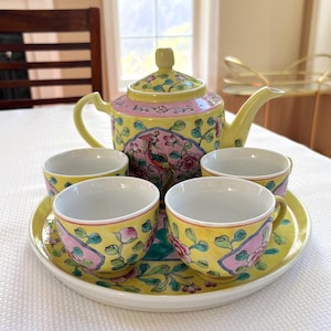 May include: A yellow ceramic tea set featuring a teapot and four matching cups on a round tray. The pieces are decorated with pink floral patterns, green leaves, and a central bird illustration on the teapot. The interior of each cup is white.