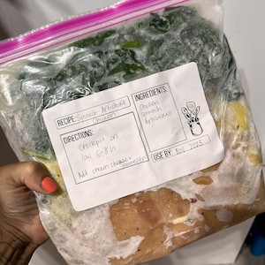 May include: A clear plastic freezer bag containing frozen chicken, spinach, and artichoke hearts. The bag has a handwritten label that reads "RECIPE: Spinach Artichoke Chicken" and "INGREDIENTS: Chicken Spinach Artichokes". The label also includes directions for cooking the ingredients in a slow cooker and a use-by date of November 2023.