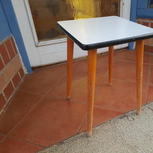 Könnte beinhalten: Ein weißer Tisch mit schwarzem Rand und vier Holzbeinen. Der Tisch steht auf einem Fliesenboden.