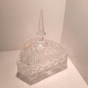 Könnte beinhalten: Transparente Bonbonniere aus geschliffenem Glas mit Deckel und spitzem Knauf. Die Schale hat einen rechteckigen Boden mit Rautenmuster und ein Blumendesign auf dem Deckel. Auf der Seite ist ein kleiner Aufkleber angebracht.