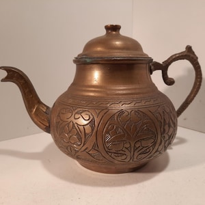 Könnte beinhalten: Eine kupferfarbene Teekanne mit Deckel und gebogenem Ausguss. Die Teekanne weist auf ihrer Oberfläche aufwendige Blumen- und Blattmuster auf. Der Griff ist geschwungen und verziert.