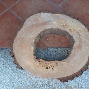 Könnte beinhalten: Ein natürlicher Holzscheibe mit einem großen, kreisförmigen Loch in der Mitte. Das Holz hat eine hellbraune Farbe mit einem dunklen, strukturierten Rindenrand. Die Holzscheibe liegt auf einer grau-braunen Fliesenoberfläche.