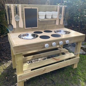 May include: Wooden play kitchen with a black chalkboard, a stainless steel sink, and a stovetop with four burners. The kitchen has a shelf for storage and hooks for hanging utensils.