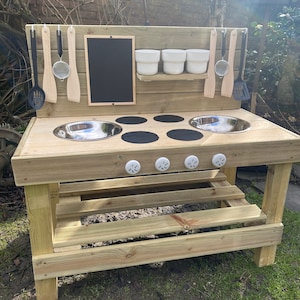 May include: Wooden outdoor play kitchen with a black chalkboard, two stainless steel sinks, a black stovetop with four burners, and a small shelf with three white bowls. The kitchen has a bench underneath and is made of natural wood.