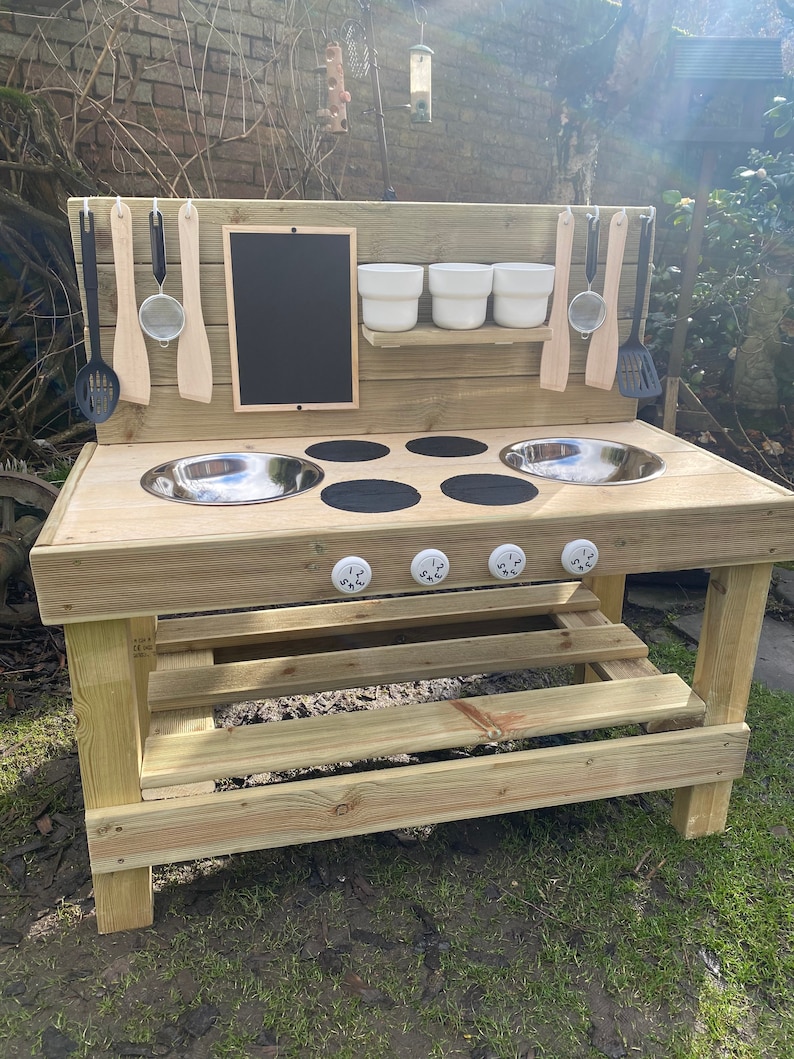 May include: Wooden outdoor play kitchen with a black chalkboard, two stainless steel sinks, a black stovetop with four burners, and a small shelf with three white bowls. The kitchen has a bench underneath and is made of natural wood.