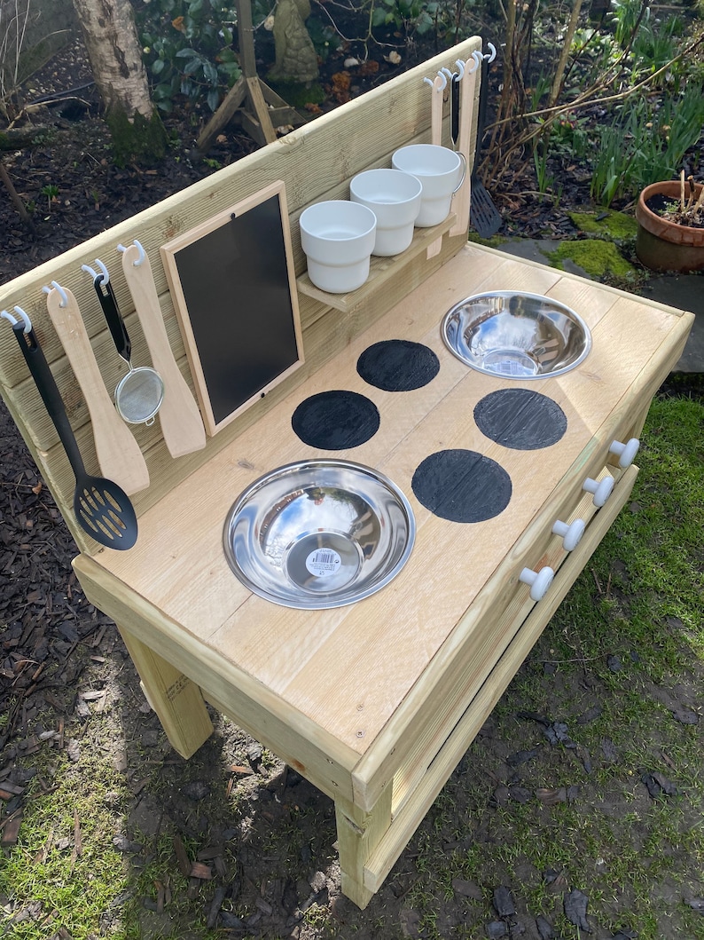 May include: Wooden play kitchen with a black chalkboard, a shelf with white bowls, and two stainless steel sinks. The kitchen has four black stovetop burners and white knobs.