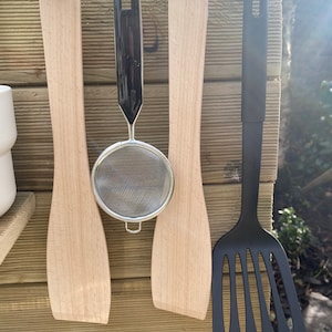 May include: Four kitchen utensils hanging on hooks. Two wooden spatulas, a black mesh strainer, and a black spatula with holes.