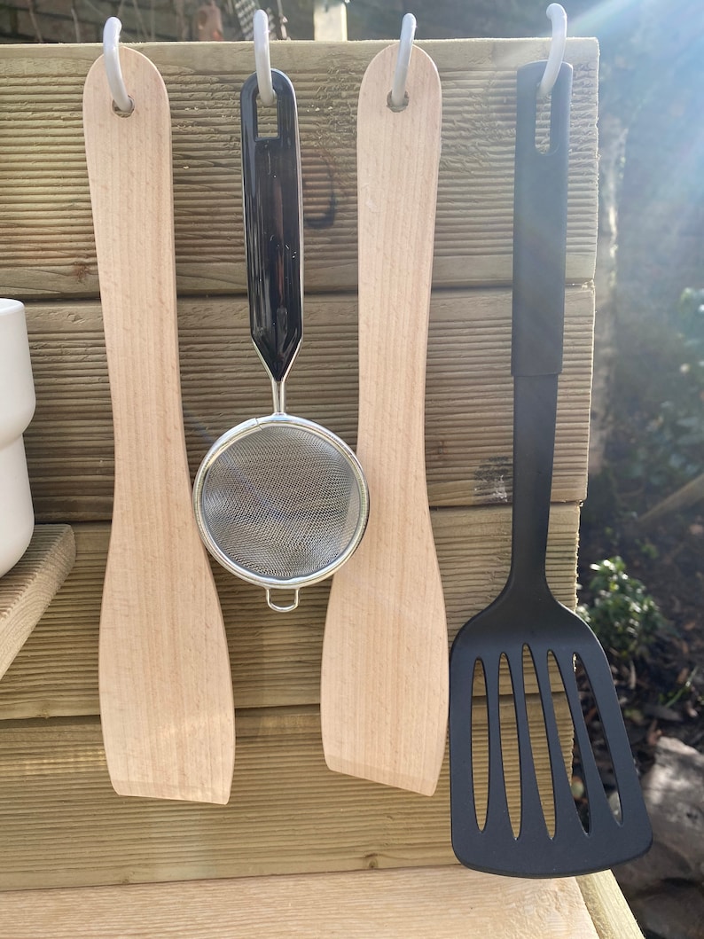 May include: Four kitchen utensils hanging on hooks. Two wooden spatulas, a black mesh strainer, and a black spatula with holes.